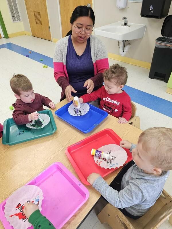 A teacher assists three young children with a gluing activity at a classroom table. The children are focused on gluing objects onto paper placed on trays.